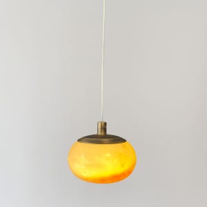 Puede incluir: Una lámpara colgante con una pantalla de vidrio amarilla y redonda y una tapa de metal color latón. La luz cuelga de un cable blanco. La pantalla de vidrio emite un brillo cálido y acogedor.