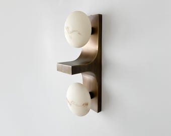 May include: A wall-mounted light fixture featuring a vertical, dark bronze-toned metal frame with a central horizontal shelf. Two smooth, oval-shaped white glass globes with subtle grey veining are attached to the frame, one above and one below.