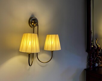 May include: A gold wall-mounted light fixture with two cream-coloured lampshades. The fixture has a curved design with a single bulb in each shade.