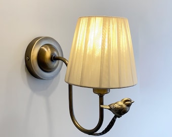 May include: A wall-mounted light fixture featuring a circular antique brass base and a curved arm. A small, detailed metal bird figure is perched on the arm, supporting a pleated, off-white fabric lampshade that emits a warm glow.