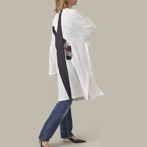 Könnte beinhalten: Eine weiße Langarmtunika mit einem schwarzen vertikalen Streifen und einem grafischen Design auf dem Rücken. Die Tunika wird mit blauen Jeans und schwarzen Absätzen getragen. Die Tunika hat einen Schlitz.