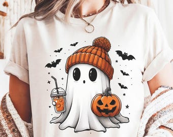 May include: Cream-colored t-shirt featuring a cartoon ghost wearing an orange knit cap. The ghost holds a pumpkin and a drink with a straw. Black bats and stars are scattered around the ghost, creating a Halloween theme.