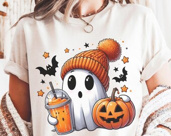 May include: Cream-colored t-shirt featuring a cartoon ghost wearing an orange knit cap with a pom-pom. The ghost holds an orange drink and is next to a carved pumpkin. Black bats and orange stars surround the ghost, creating a Halloween theme.