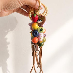 Peut inclure: Un porte-clés composé d'un anneau en métal doré attaché à une collection de perles multicolores de formes variées, incluant une fraise rouge, un cœur marron avec un œil, un cube vert avec une étoile et plusieurs perles rondes fruitées, terminées par des pompons en cuir marron.