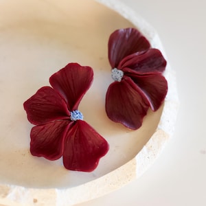 May include: A pair of deep burgundy flower-shaped earrings featuring textured, layered petals and a small, light blue speckled center. The petals have a smooth, slightly glossy finish with visible veins, creating a realistic floral appearance.