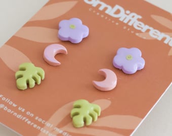 Puede incluir: Un conjunto de pendientes de resina coloridos con forma de flores, lunas y hojas. Los pendientes se muestran en una tarjeta de color melocotón con el texto "Born Different Creative Studio".