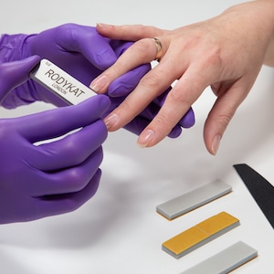 May include: A person wearing purple gloves is filing a fingernail with a silver nail file that says "RODYKAT LONDON". There are three nail files on a white surface, one is grey, one is gold, and one is black.