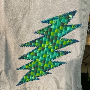 May include: A light blue pair of shorts with a green and blue lightning bolt design. The lightning bolt is made of a plaid fabric and is sewn on with a zig-zag stitch.