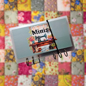 May include: A clear binder with a decorative cover featuring the word "Minis" above an illustration of a sewing machine on a table. The cover has a light blue background with white polka dots. A black elastic band holds the binder closed.