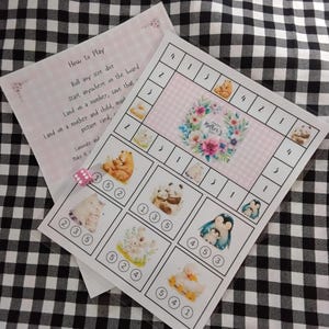 Könnte beinhalten: Ein rosa-weißes Spielbrett mit einem floralen Kranz in der Mitte, auf dem "Happy Mother's Day" steht. Das Brett hat die Zahlen 1-5 am Rand und Bilder von Tieren mit ihren Müttern in der Mitte. Zum Spiel gehört ein rosa Würfel mit weißen Punkten.