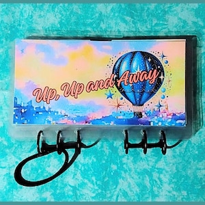 May include: A key holder with a colorful design featuring a blue hot air balloon and the words "Up, Up and Away." The key holder has a clear plastic frame and black metal hooks.