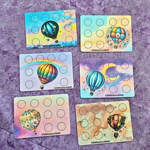 Puede incluir: Seis tarjetas rectangulares con diseños de globos aerostáticos coloridos. Cada tarjeta tiene 12 pequeños círculos. Las tarjetas presentan varios fondos, incluyendo un arcoíris, un horizonte urbano y efectos de acuarela. El texto "DSMStation5849" está impreso en cada tarjeta.