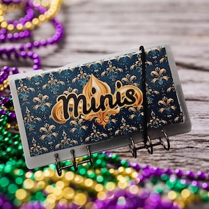 May include: A small, rectangular notebook with a decorative cover featuring a blue and gold fleur-de-lis pattern. The word "Minis" is written in gold script. The notebook is held together with a black elastic band and metal rings.
