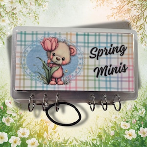 May include: A rectangular clear plastic case with a plaid background featuring a cartoon bear holding a pink tulip inside an oval frame. The text Spring Minis is printed in black on the right side. A black elastic hair tie hangs from the metal rings.