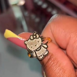 May include: A gold-toned ring featuring a flat, textured charm shaped like the character Hello Kitty. The charm displays the iconic cat face with a bow on one side, whiskers, and a simple dress design, all rendered in a metallic finish with a grid pattern.