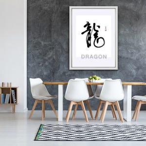May include: A framed print featuring a black and white calligraphy design of the Chinese character for "dragon", with the word "Dragon" printed below in black text. The print is hanging on a grey wall above a wooden table with white chairs.
