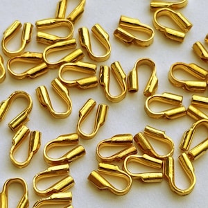 May include: A group of gold-colored metal jewelry findings, also known as jump rings, are scattered on a white surface. The jump rings are small and have a rounded shape.