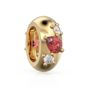 Könnte beinhalten: Ein goldfarbener Perlenanhänger mit poliertem Finish. Der Anhänger hat einen großen, birnenförmigen roten Edelstein und zwei kleinere weiße Edelsteine. Im Inneren der Perle ist ein rosa herzförmiger Akzent zu sehen.