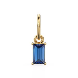 May include: A gold charm with a blue rectangular gemstone. The charm has a loop at the top for attaching to a necklace or bracelet.