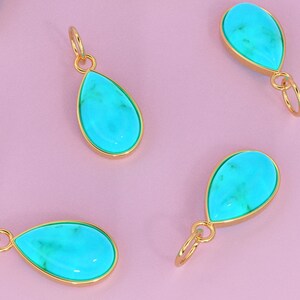 May include: Seven turquoise teardrop-shaped pendants with gold settings on a pink background. The pendants are all the same size and shape, and they are arranged in a random pattern.