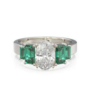 Puede incluir: Un anillo de tono plateado con una gran piedra preciosa central transparente de corte ovalado, flanqueada por dos piedras verdes de corte esmeralda a cada lado. La banda es lisa y pulida, sujetando las cinco piedras en un engaste.