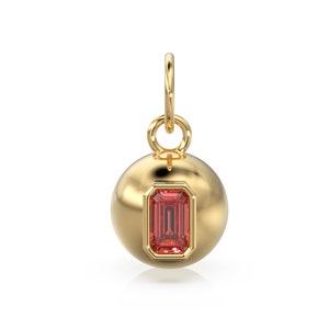 May include: A gold charm with a red gemstone in the shape of an emerald cut. The charm is in the shape of a sphere with a loop at the top for attaching to a necklace or bracelet.
