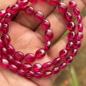 May include: A strand of oval-shaped ruby beads in a deep red color. The beads are strung on a wire and are arranged in a circular pattern.