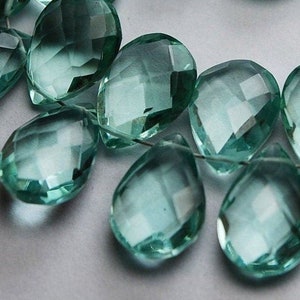 May include: A strand of faceted teardrop-shaped beads in a light aqua blue color. The beads are strung on a clear thread and are arranged in a circular pattern.