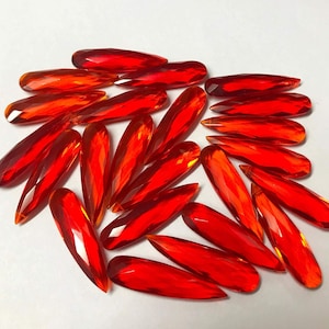 Peut inclure: Une collection de perles facettées en forme de goutte d'eau, d'une couleur rouge translucide et vibrante. Chaque perle présente de multiples surfaces polies qui captent la lumière, créant un aspect brillant semblable à une pierre précieuse.