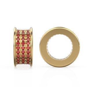 Puede incluir: Dos anillos dorados. Uno se muestra de lado, revelando una banda de pequeñas piedras preciosas rojas cuadradas. El otro anillo se muestra de frente, mostrando una banda dorada con un interior blanco.