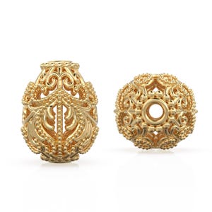 May include: Two gold-colored filigree beads. One is oval-shaped with ornate openwork and a beaded texture. The other is round with a central hole and a similar openwork design. Both beads have a detailed, vintage aesthetic.