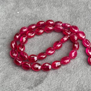 May include: A strand of oval-shaped, bright red ruby beads. The beads are strung together on a clear thread.