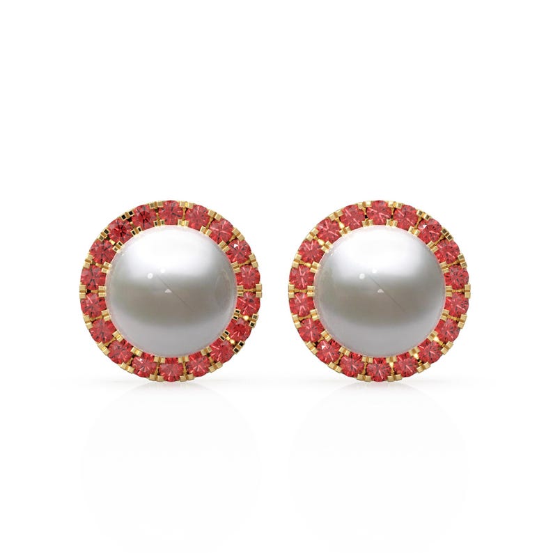 May include: A pair of stud earrings with a large, round, white pearl centre, encircled by a halo of small, red gemstones. The setting is gold-coloured, contrasting with the white and red elements.