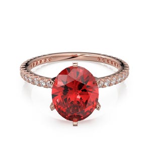 Può includere: Un anello in oro rosa con una grande pietra preziosa rosso vivo dal taglio ovale, fissata da sei griffe. La fascia è decorata con una serie di piccole pietre bianche trasparenti dal taglio rotondo incastonate lungo i lati dell'anello.