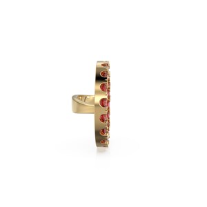 May include: A gold ring with a vertical design, set with small, round, red gemstones. The ring's band is plain, while the gemstone section has a decorative, openwork pattern. The ring is a piece of jewellery.