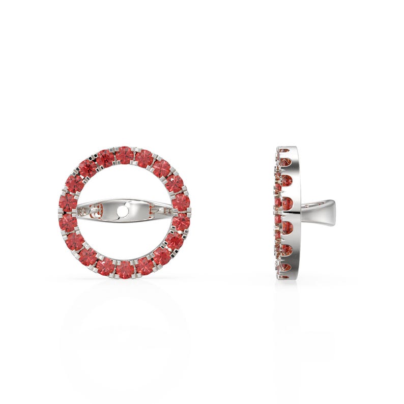 May include: A pair of silver earrings. Each earring has a circular design with a ring of small, red gemstones. The earrings are shown from the front and side, showcasing the design and the post.