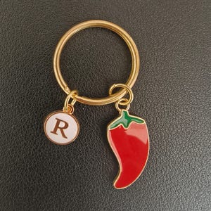May include: A gold key chain with a red and green chilli pepper charm and a white circle charm with the letter "R" on it.