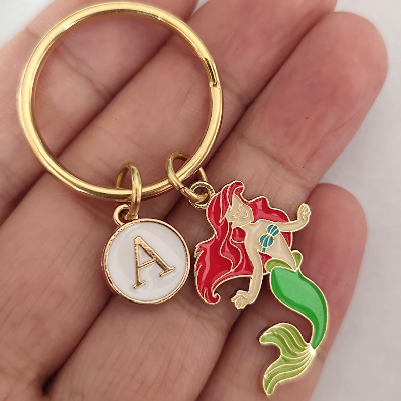 May include: Gold key chain with a white circle charm featuring the letter A and a mermaid charm with red hair and a green tail.