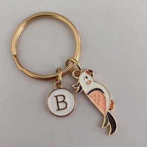 May include: Gold key ring with a white circular charm featuring the letter "B" and a pink and white bird charm.