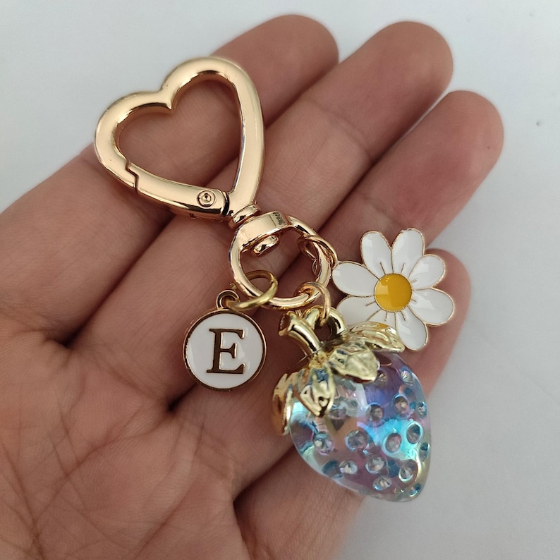 May include: Gold-tone keychain with a heart-shaped ring, a charm with the letter E, a blue and white glass strawberry charm, and a white daisy charm.