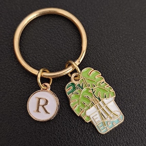 May include: Gold-tone keychain with two charms: a white circle with the letter "R" and a green and white plant design. The plant charm features a monstera plant in a white pot with a light blue base. The keychain is a decorative accessory.
