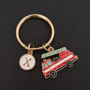 May include: A gold-coloured keyring featuring a red and white fire engine charm and a white circular charm with the letter "X". The fire engine has black wheels and a blue ladder. The keyring is on a dark surface.