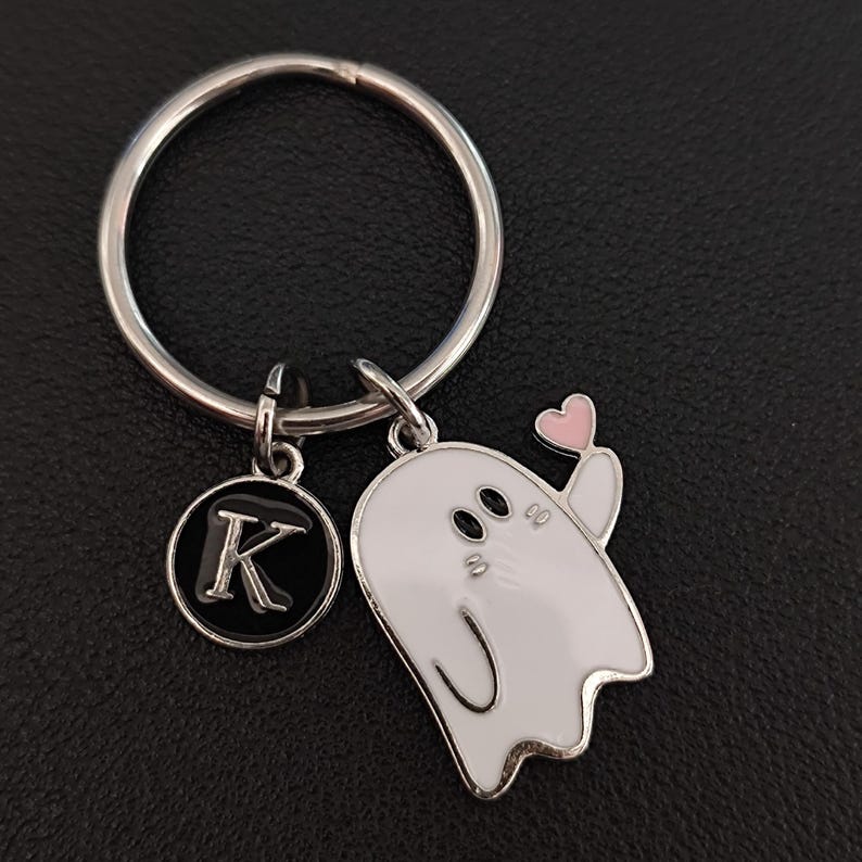 May include: A silver keychain with a round key ring. The keychain has two charms: a black and silver circular charm with the letter "K" and a white ghost charm holding a pink heart. The ghost has black eyes and a curved mouth.
