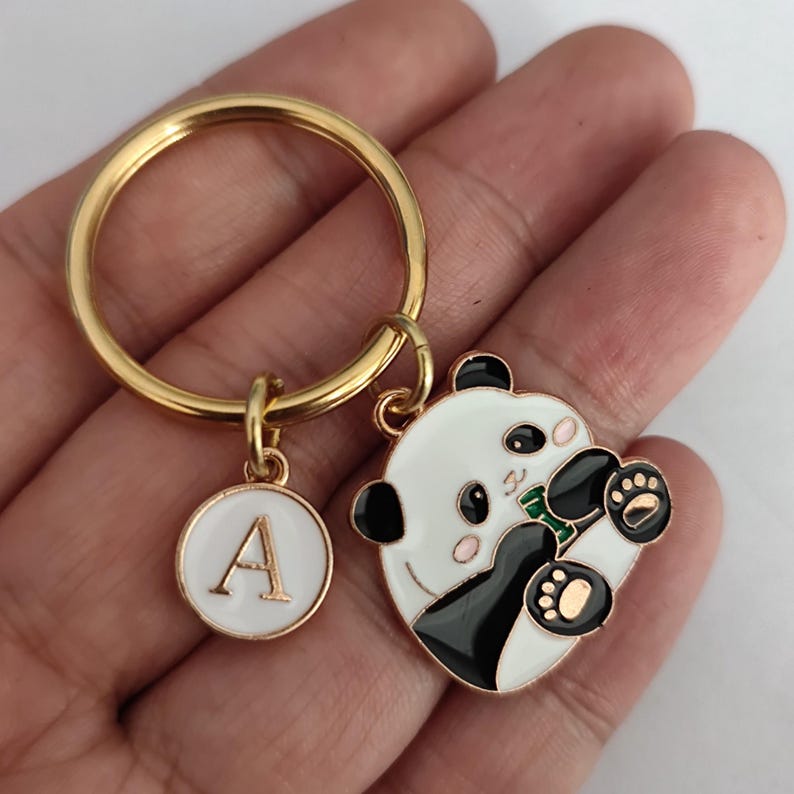 May include: Gold key chain with a black and white panda charm holding a green bamboo shoot and a white circle charm with the letter A.