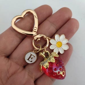 May include: Gold-tone keychain with a heart-shaped ring, a red and gold strawberry charm, a white daisy charm, and a white circle charm with the letter 'E' in gold.