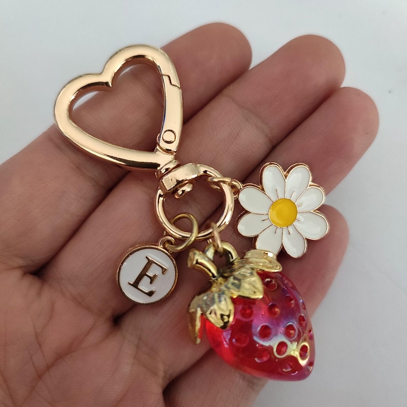 May include: Gold-tone keychain with a heart-shaped ring, a red and gold strawberry charm, a white daisy charm, and a white circle charm with the letter 'E' in gold.