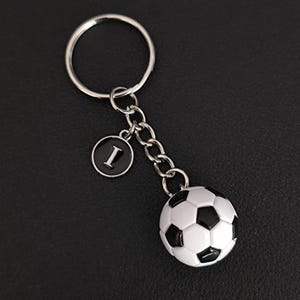 May include: A soccer ball keychain with a silver-colored ring and chain. The keychain includes a black and white soccer ball charm and a circular charm with the letter "I" in white on a black background.