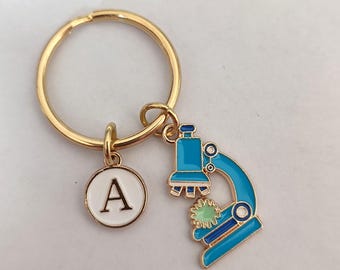 Può includere: Portachiavi dorato con un ciondolo a microscopio blu e bianco e un ciondolo a cerchio bianco con la lettera A in nero.