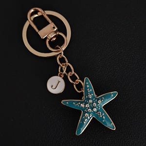 May include: A gold-colored keychain with a teal starfish charm adorned with small, clear crystals. The keychain includes a round charm with the letter "J" and a lobster clasp.
