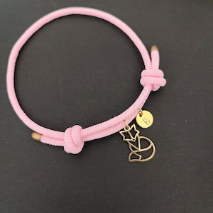 Könnte beinhalten: Ein pinkfarbenes geflochtenes Armband aus Seil mit einem goldenen Anhänger, der einen Stern und eine Fuchs-Silhouette zeigt. Der Anhänger hat auch eine kleine runde Scheibe mit dem eingravierten Buchstaben "C".